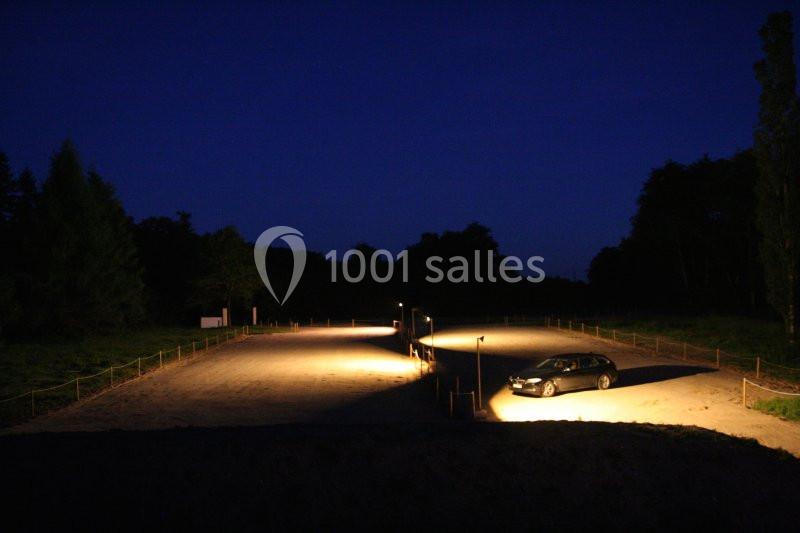 Un parking vide éclairé par des lampadaires la nuit, avec une seule voiture stationnée à droite.