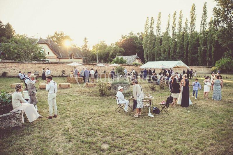 Groupe de personnes réunies dans un jardin, avec des tables, des bottes de foin et des tentes sous un ciel ensoleillé.