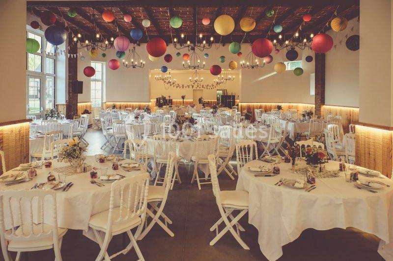 Salle décorée pour un événement, avec des tables rondes dressées, des chaises blanches et des lanternes colorées suspendues.