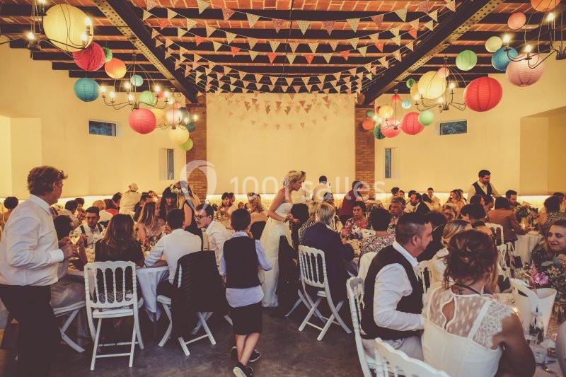 Salle décorée avec des guirlandes et lanternes colorées, remplie d'invités assis à des tables lors d'une réception.