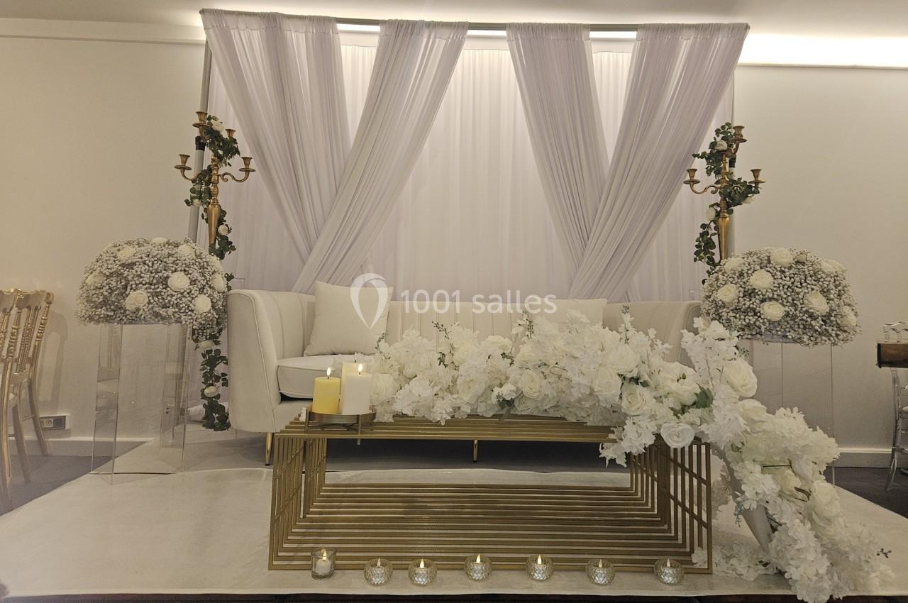 Décoration intérieure élégante avec canapé blanc, rideaux blancs, fleurs blanches et bougies sur une table dorée.