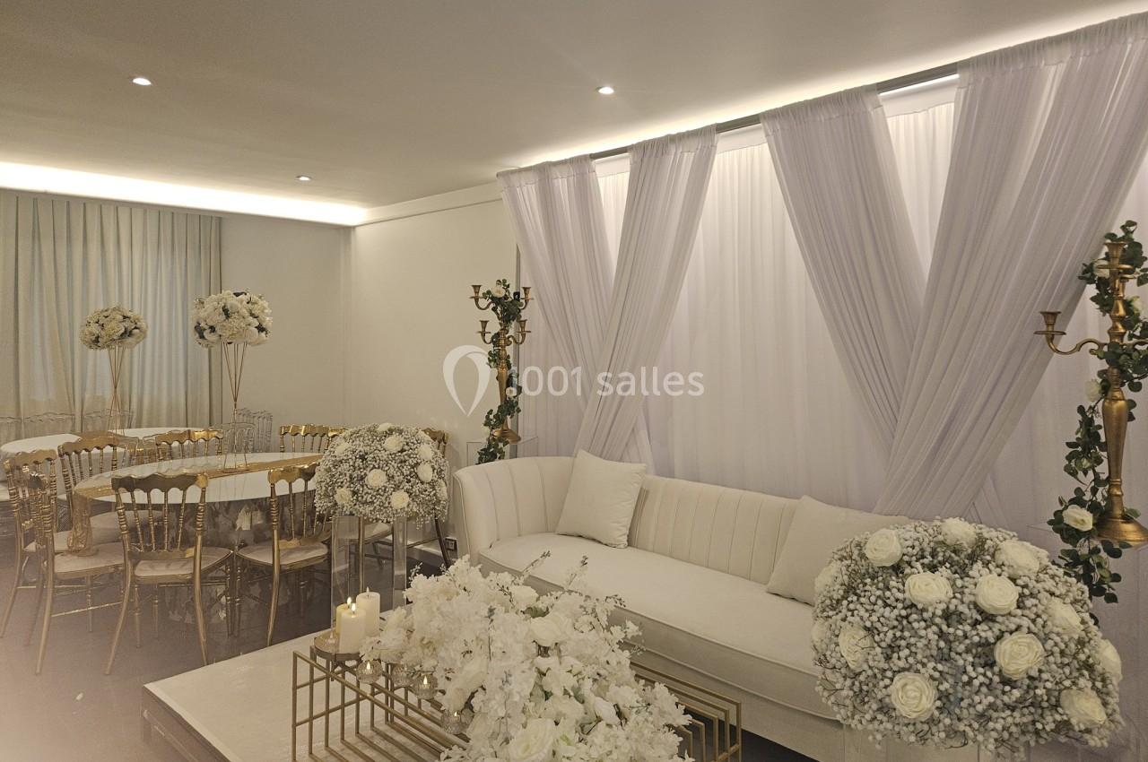 Salle décorée pour un événement avec canapés blancs, rideaux, chandeliers dorés et compositions florales blanches.