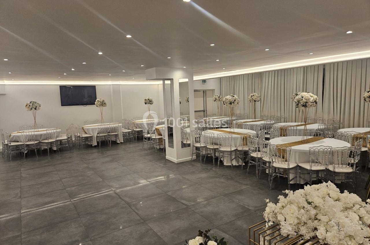 Salle de réception moderne avec tables dressées, chaises transparentes et décorations florales blanches.