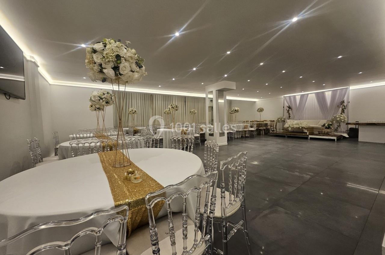Salle de réception élégante avec tables décorées de nappes dorées, chaises transparentes et arrangements floraux blancs.
