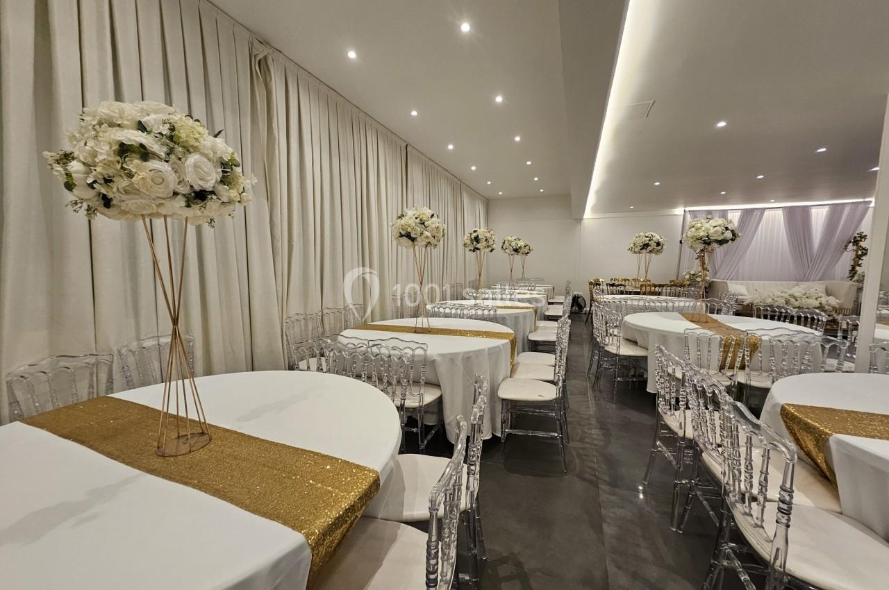 Salle de réception décorée avec des tables rondes, nappes blanches, chemins de table dorés et centres de table fleuris.