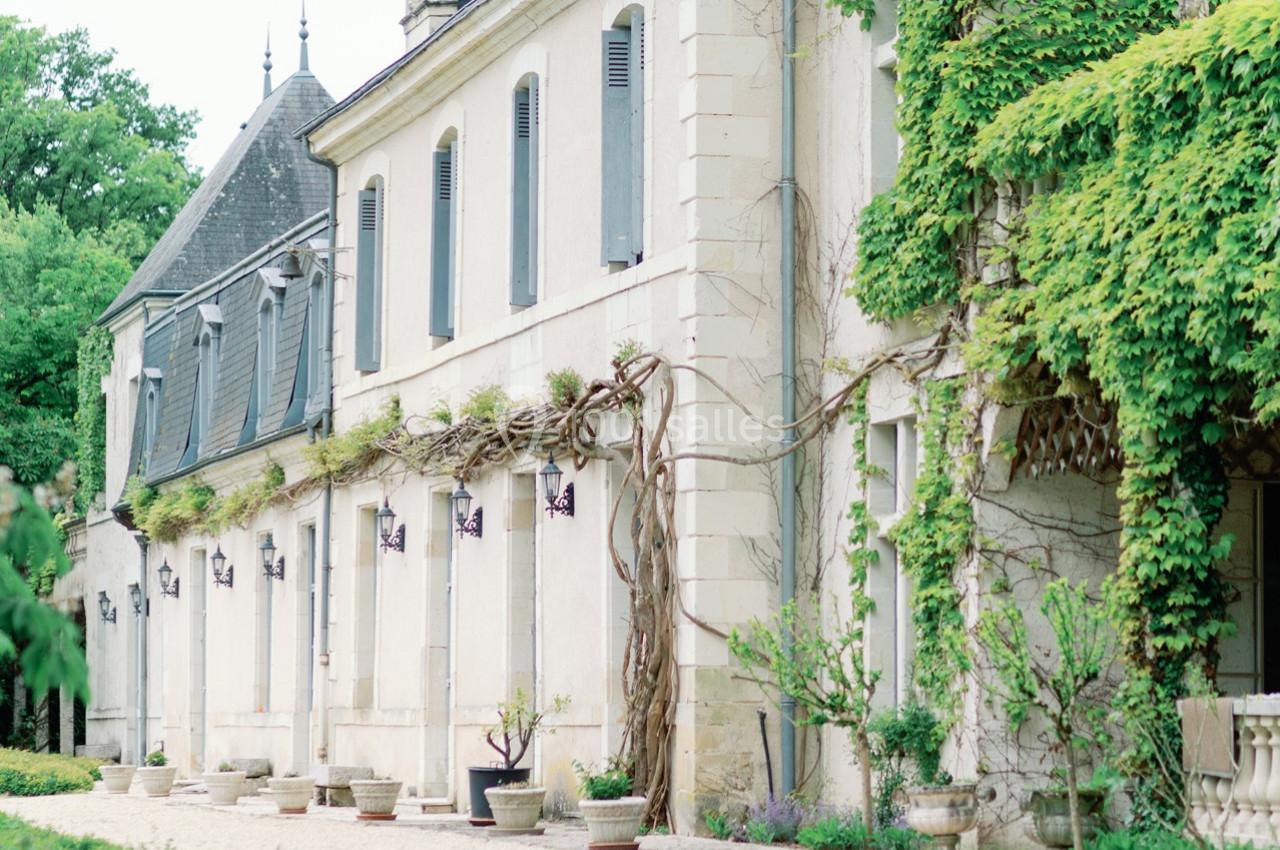 Façade d'un bâtiment ancien en pierre, partiellement recouverte de vigne, avec des pots de fleurs devant.
