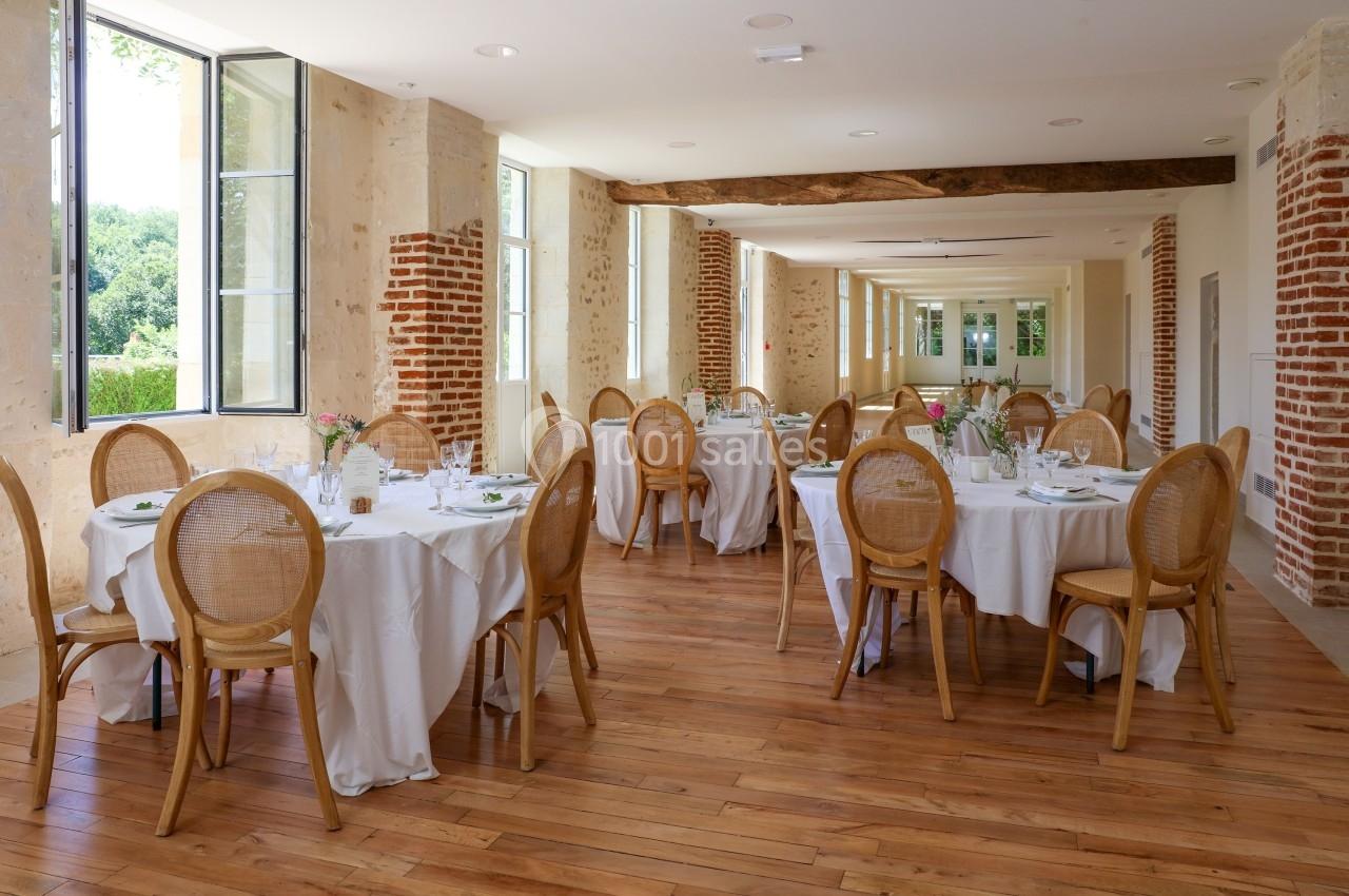 Salle lumineuse avec des tables rondes dressées, chaises en bois clair et grandes fenêtres ouvertes sur un jardin.