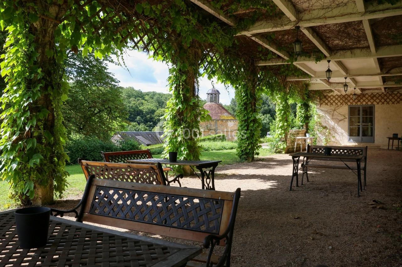 Terrasse ombragée avec tables en fer forgé, entourée de verdure et vue sur un bâtiment ancien dans un cadre rural.