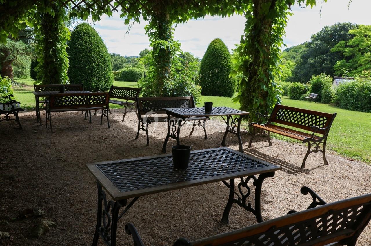 Tables et bancs en fer forgé sous une pergola ombragée, entourés de verdure dans un jardin.