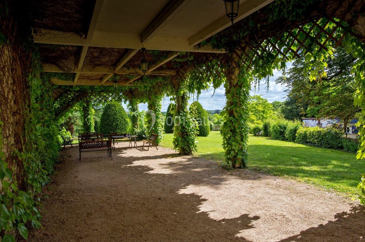 Allée ombragée sous une pergola couverte de végétation, avec des bancs et une table, donnant sur un jardin verdoyant.