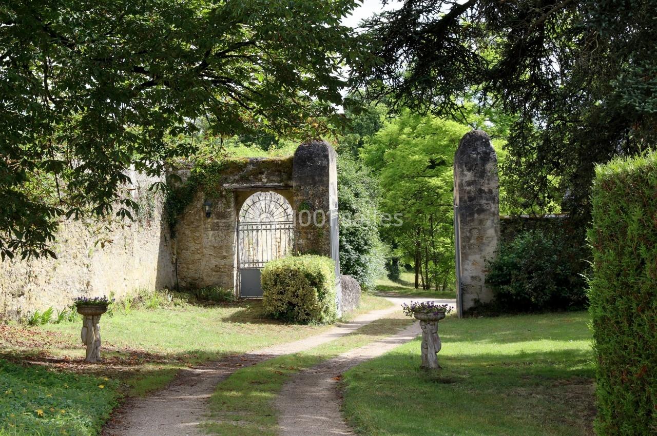 Allée bordée de verdure menant à une porte en fer forgé encadrée par des piliers en pierre dans un jardin.