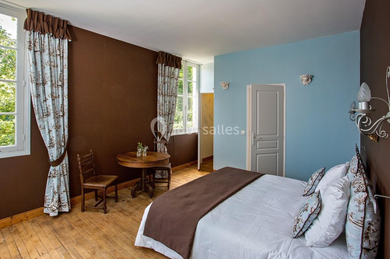 Chambre lumineuse avec murs bleu et marron, lit double, table en bois, chaises et fenêtres donnant sur la verdure.