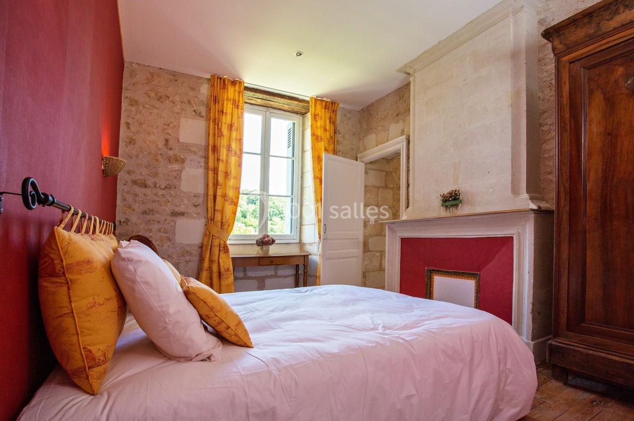 Chambre lumineuse avec murs en pierre, cheminée blanche, rideaux jaunes et lit avec coussins assortis.