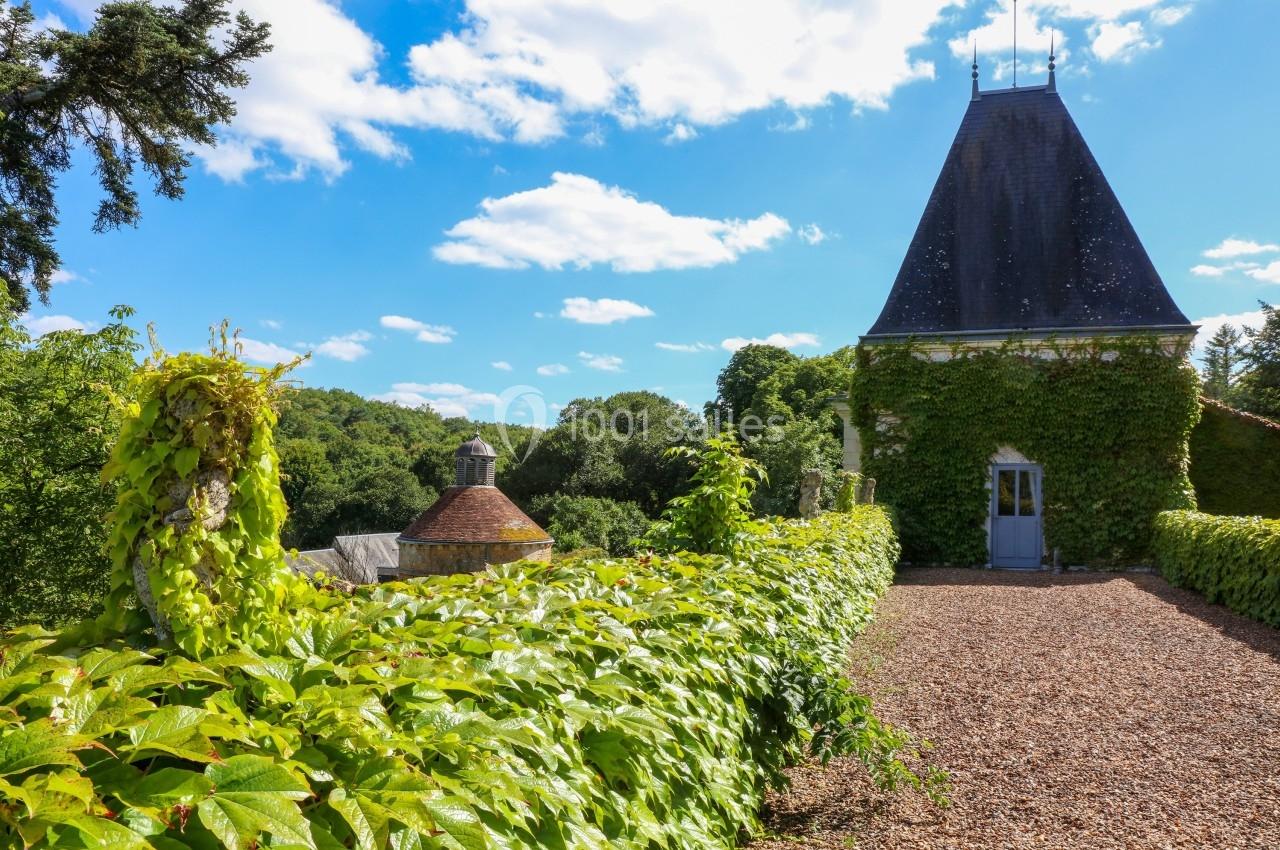 Allée bordée de verdure menant à une tour couverte de lierre dans un paysage rural sous un ciel bleu.