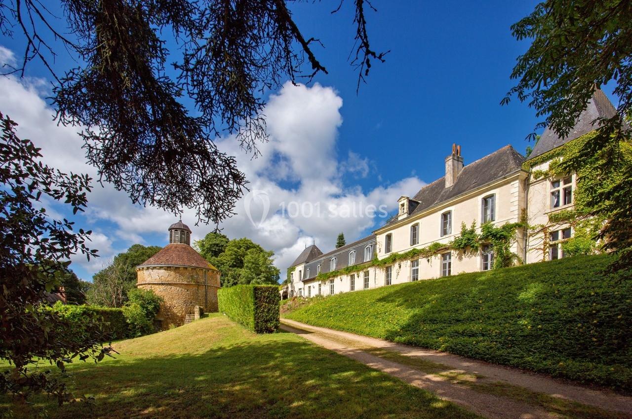 Manoir en pierre avec un colombier rond, entouré de verdure sous un ciel bleu parsemé de nuages.