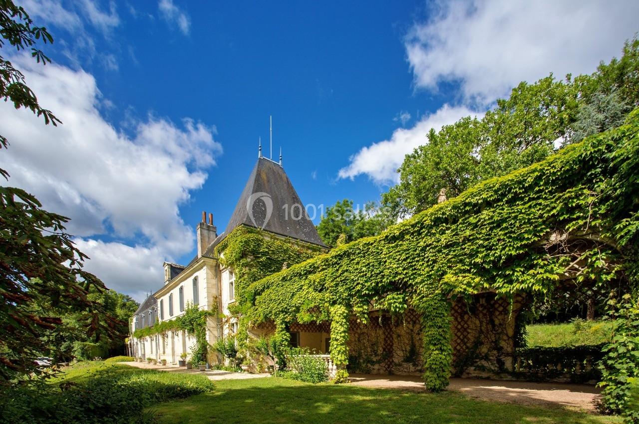 Façade d'une maison ancienne recouverte de lierre, entourée de verdure sous un ciel bleu partiellement nuageux.