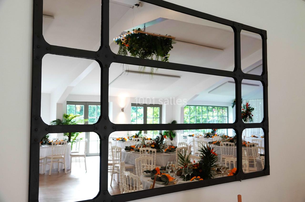 Reflet d'une salle de réception décorée avec des tables dressées et des plantes, visible dans un grand miroir à cadres noirs.