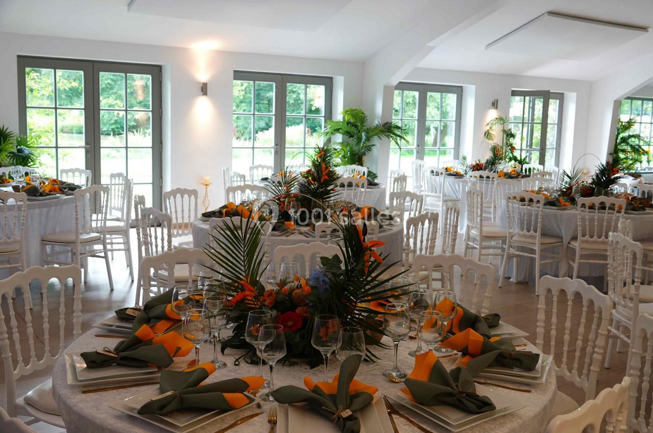 Salle de réception lumineuse avec tables décorées de nappes blanches, serviettes orange et arrangements floraux tropicaux.