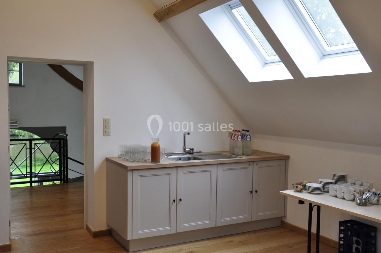 Petite kitchenette lumineuse avec évier, bouteilles d'eau, verres et vaisselle, sous un toit en pente avec fenêtres.