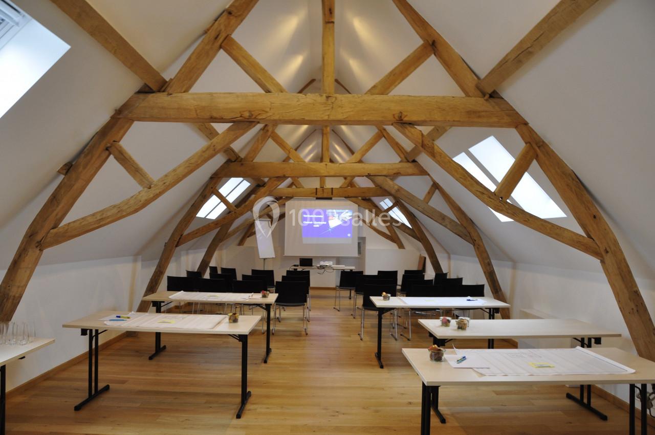 Salle de réunion lumineuse sous combles avec poutres apparentes, tables, chaises et écran de projection installé.