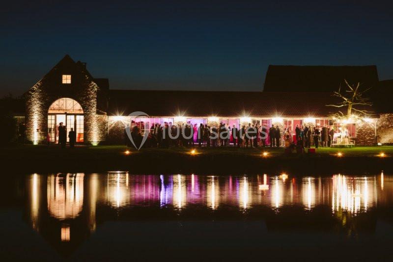 Bâtiment éclairé en soirée avec des invités rassemblés à l'extérieur, reflété dans un plan d'eau calme.