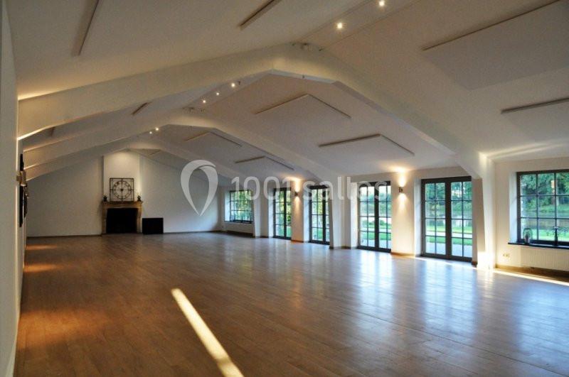 Salle spacieuse avec parquet en bois, grandes fenêtres laissant entrer la lumière naturelle, et cheminée au fond.