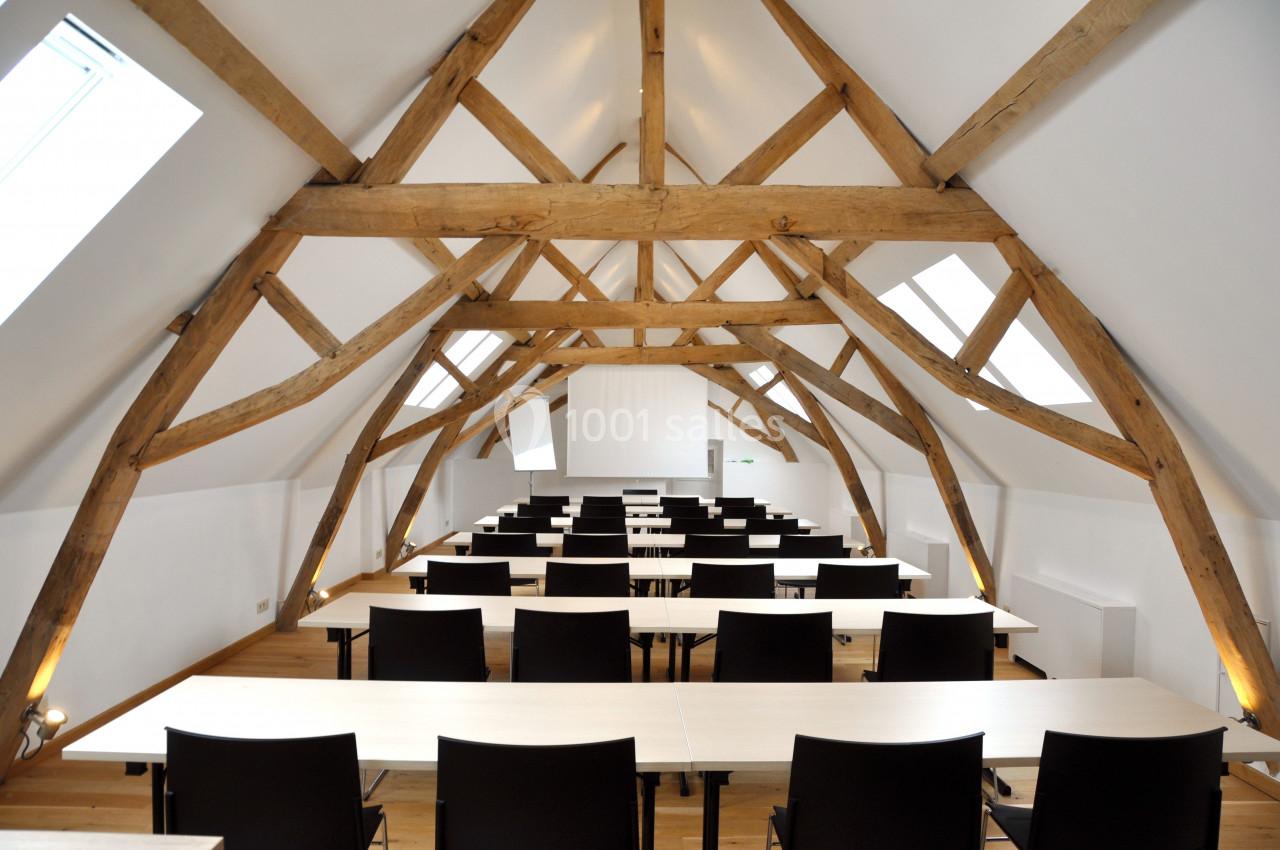Salle de formation lumineuse sous combles avec poutres apparentes, équipée de tables et chaises alignées.