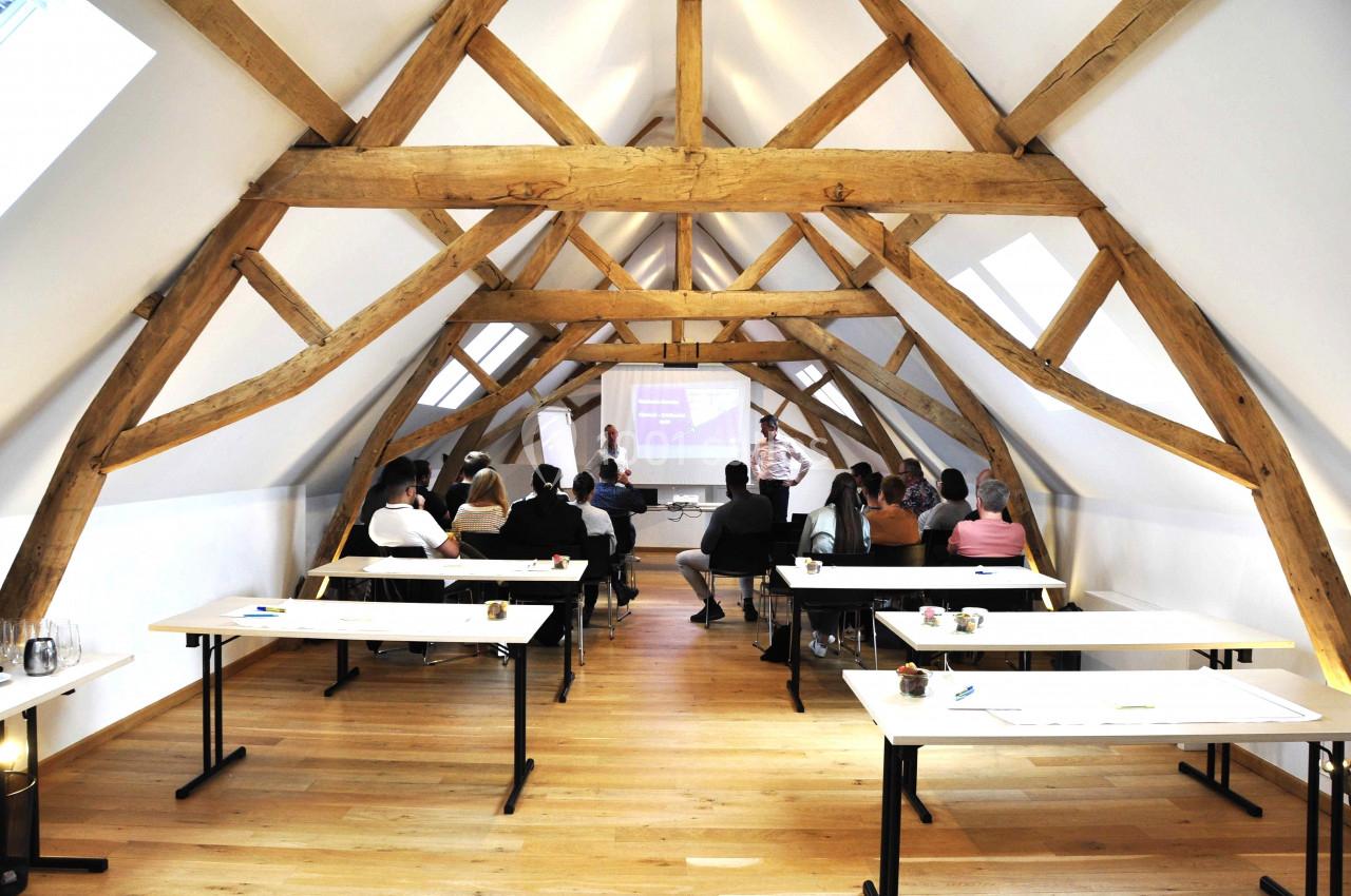 Salle de réunion sous combles avec poutres apparentes, où un groupe de personnes assiste à une présentation.