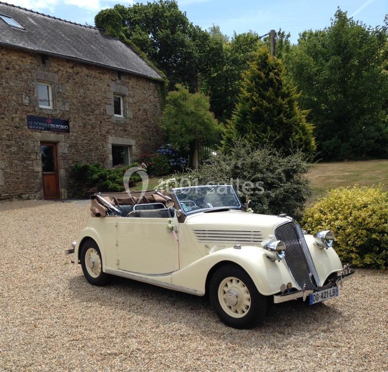 Voiture ancienne décapotable blanche stationnée sur une allée gravillonnée devant une maison en pierre et un jardin…