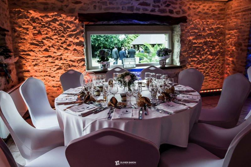 Table ronde élégamment dressée avec nappes blanches, verres et couverts, dans une salle en pierre éclairée chaleureusement.