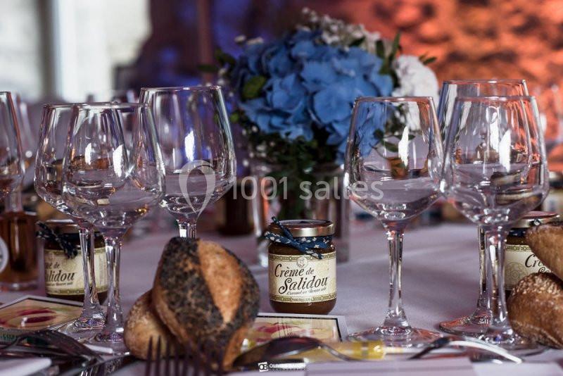 Table dressée avec des verres à pied, des pots de crème de salidou, des pains et un bouquet de fleurs bleues en arrière-plan.