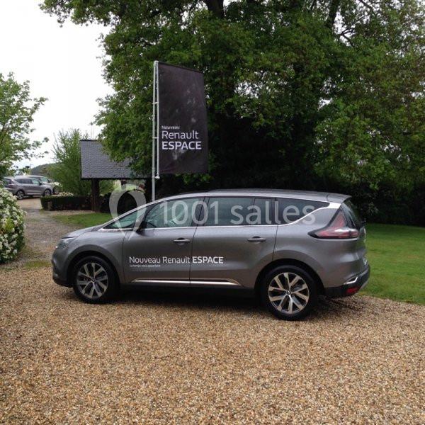 Voiture grise Renault Espace stationnée sur une allée en gravier, entourée de verdure et d'un panneau publicitaire.