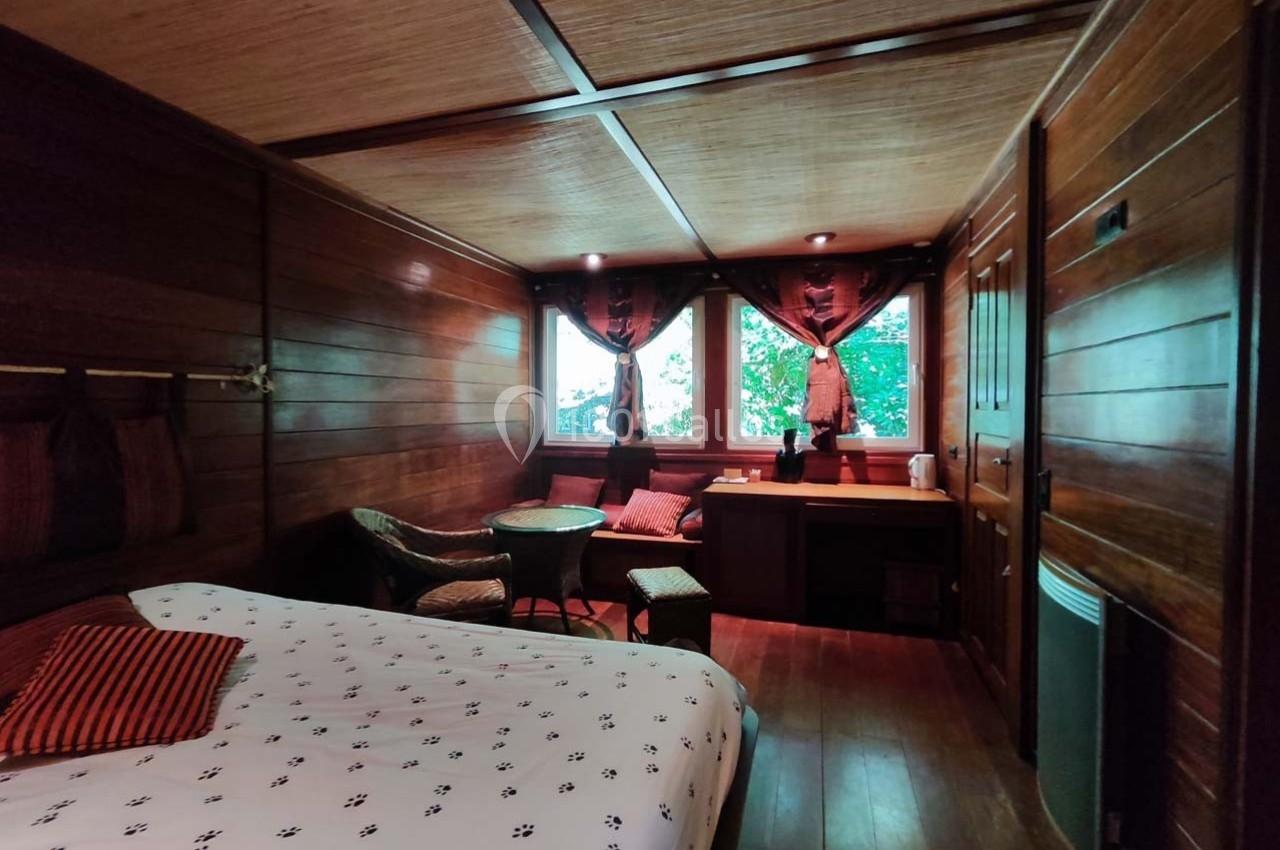 Chambre en bois avec lit, table, chaises en osier, fenêtres donnant sur un extérieur verdoyant et rideaux sombres.