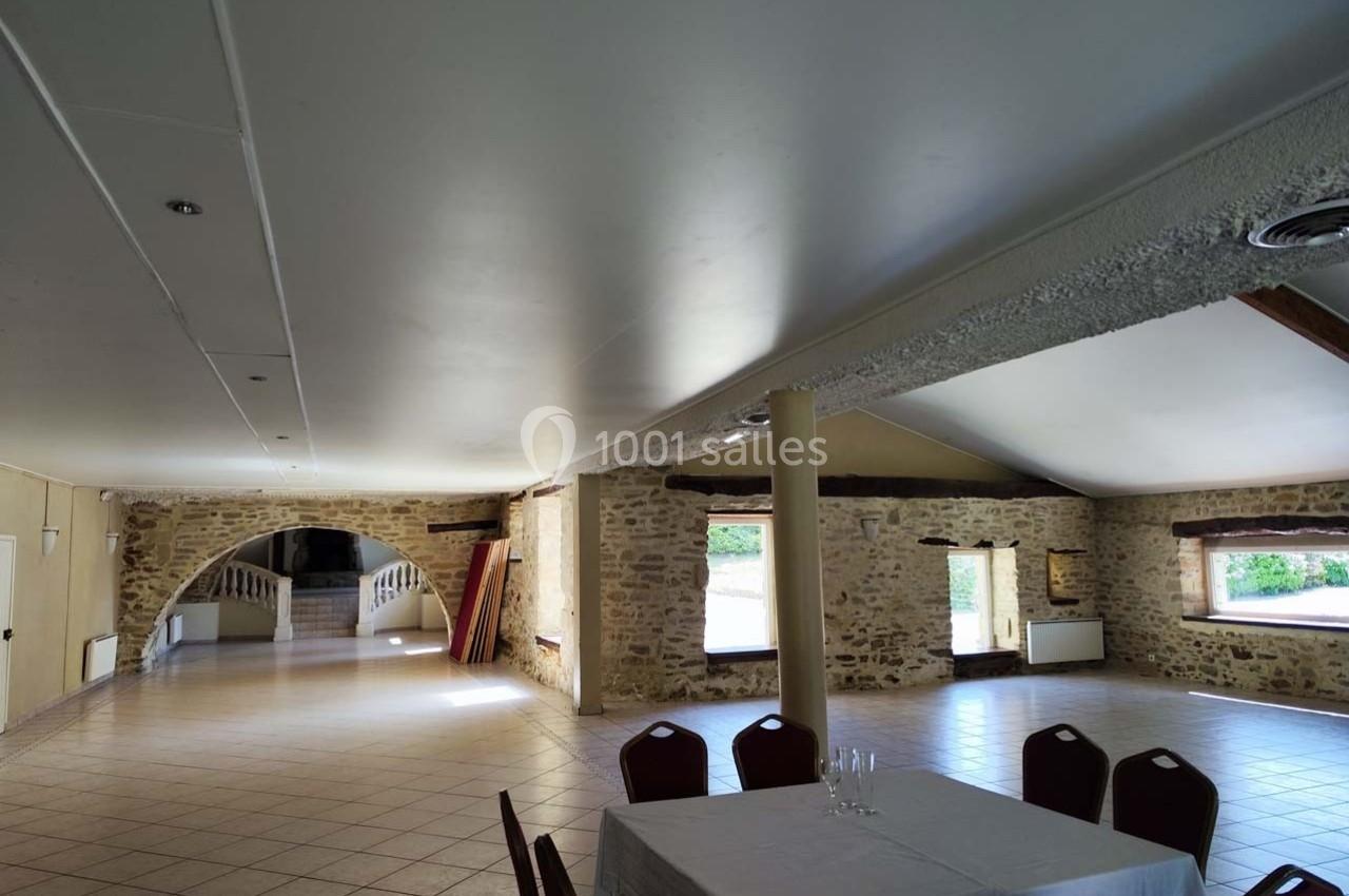 Salle spacieuse avec murs en pierre, plafond blanc, grandes fenêtres et une table dressée au premier plan.