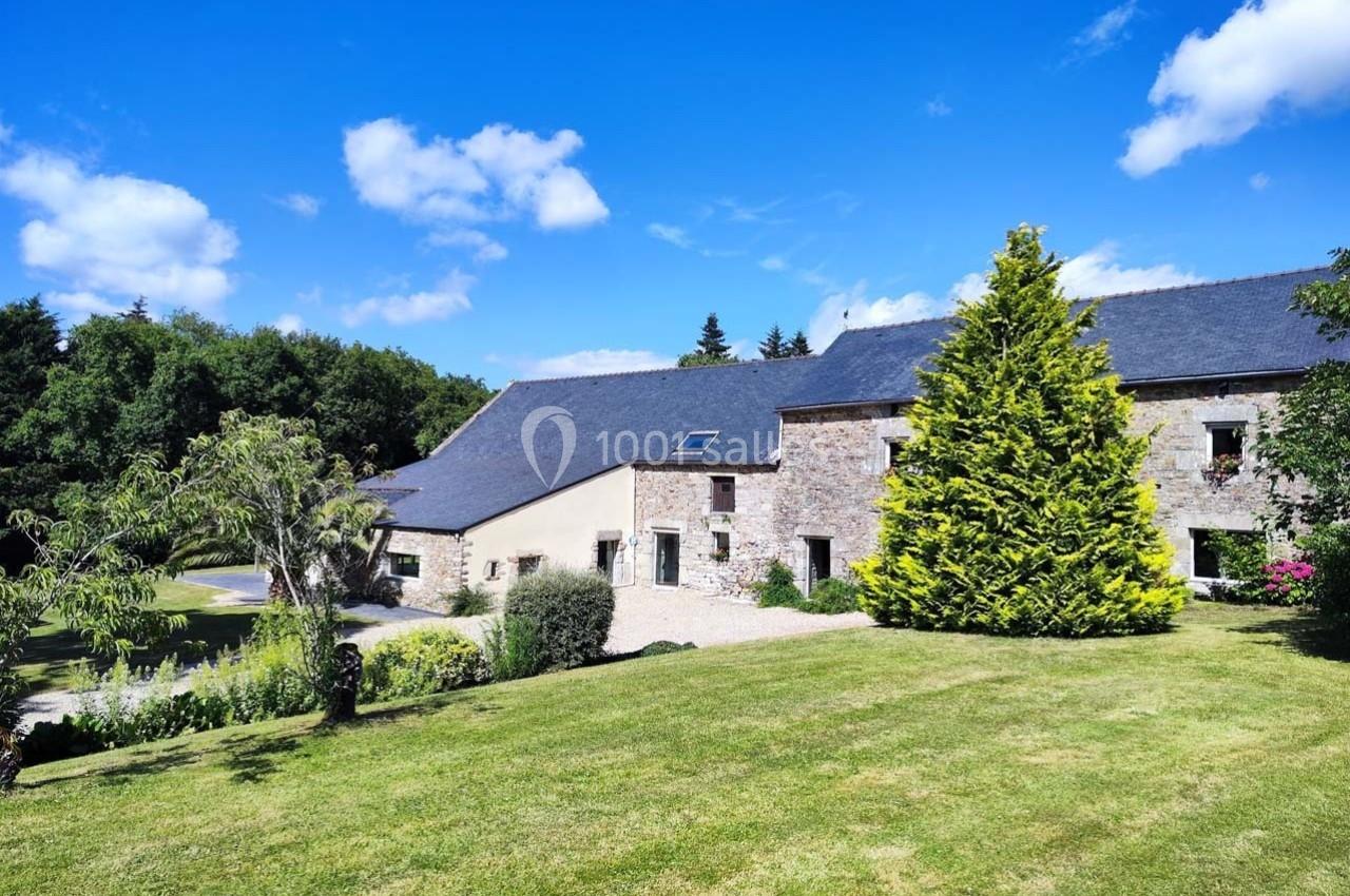 Bâtisse en pierre avec toit en ardoise entourée de verdure et d'un jardin sous un ciel bleu.