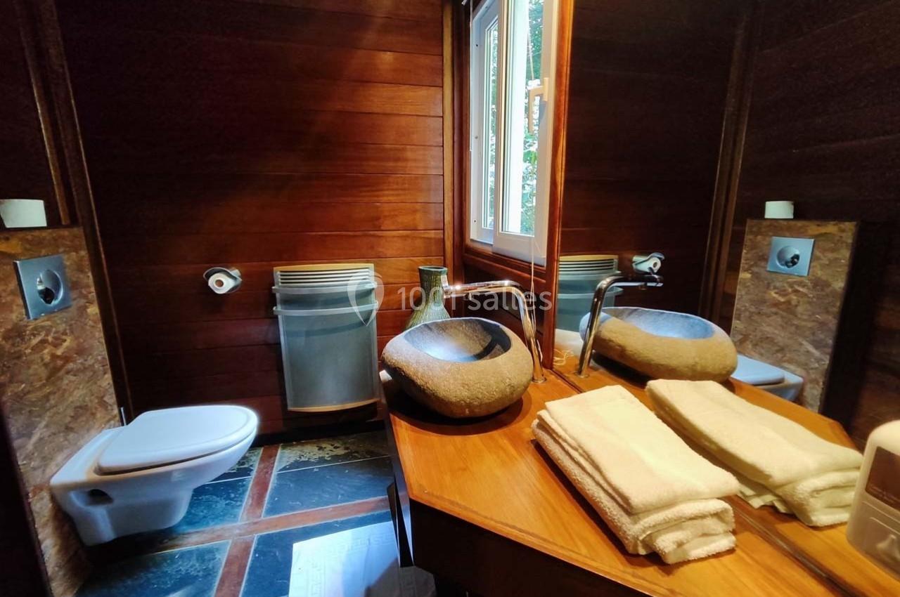 Salle de bain avec murs en bois, lavabo en pierre, miroir, toilettes suspendues et serviettes pliées sur un plan de travail.