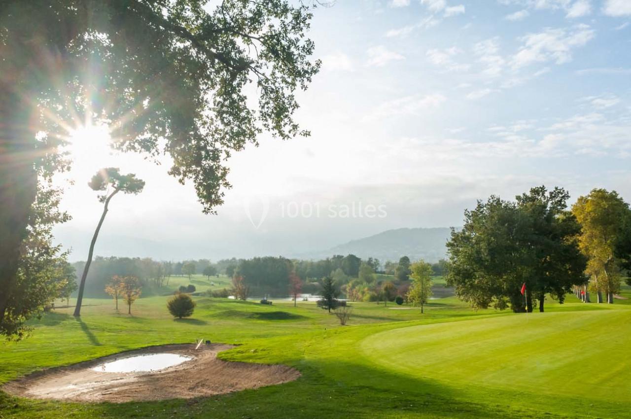 Paysage de golf avec un green, un bunker, des arbres et un soleil traversant les branches sous un ciel partiellement nuageux.