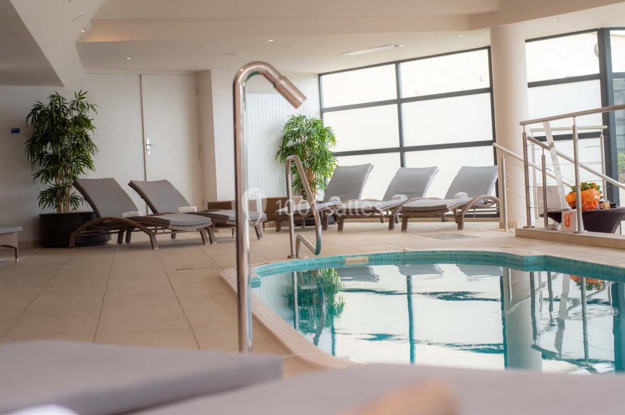 Piscine intérieure avec transats alignés, plantes décoratives et grandes baies vitrées laissant entrer la lumière naturelle.