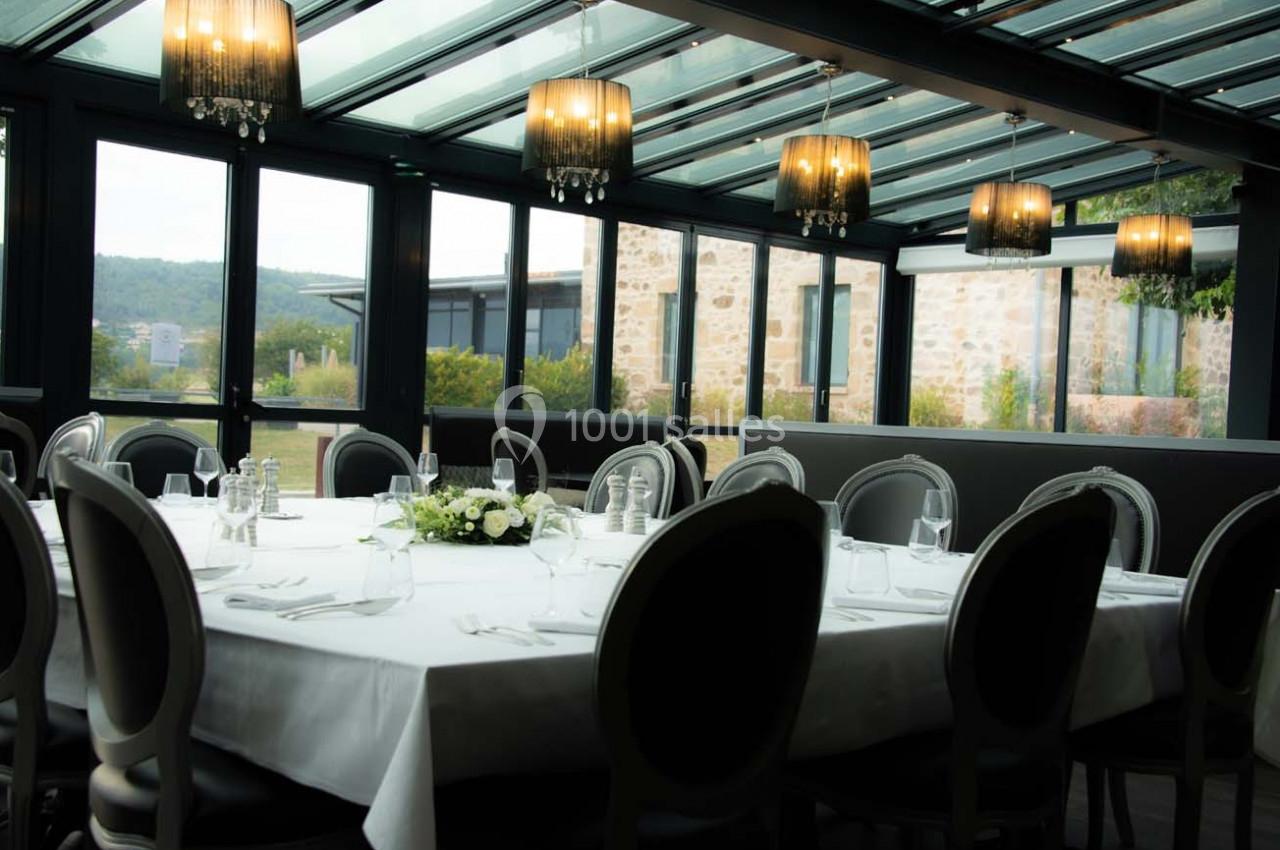 Salle à manger lumineuse avec une grande table ronde dressée, entourée de chaises élégantes, sous une verrière.