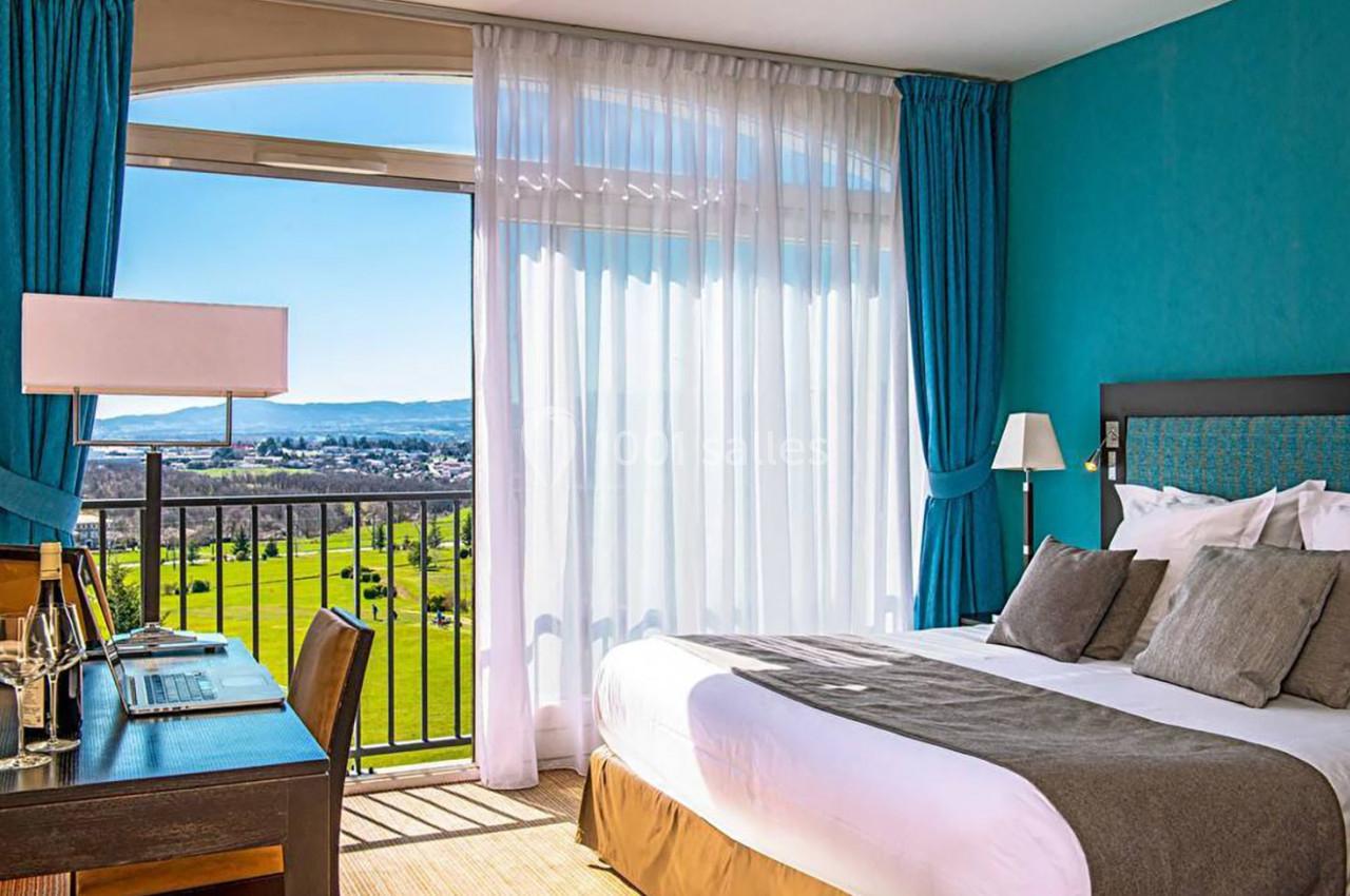 Chambre lumineuse avec lit double, bureau, et grande baie vitrée donnant sur un paysage verdoyant et ensoleillé.