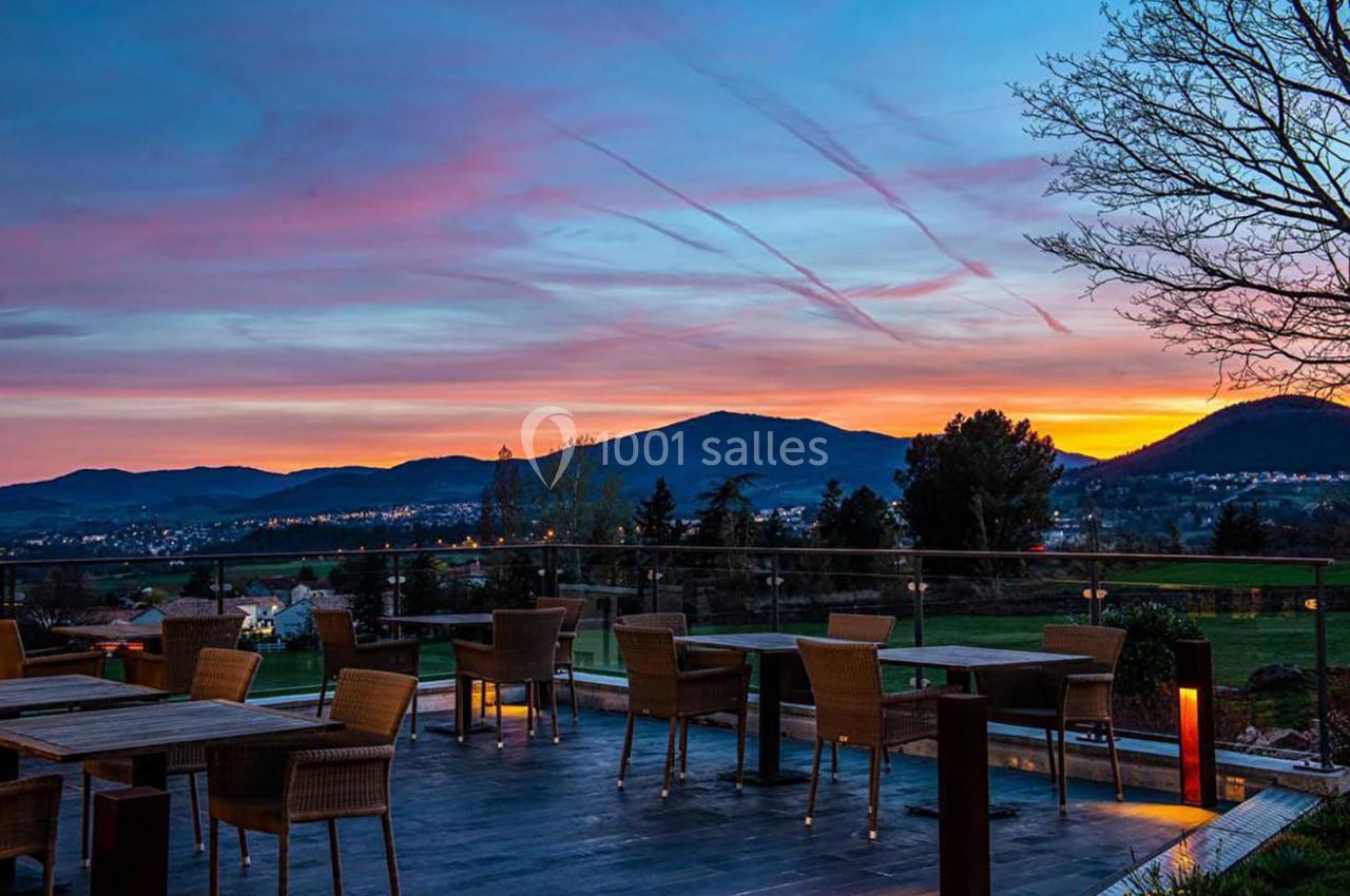 Terrasse avec des tables et chaises en rotin, offrant une vue sur un paysage vallonné au coucher du soleil.
