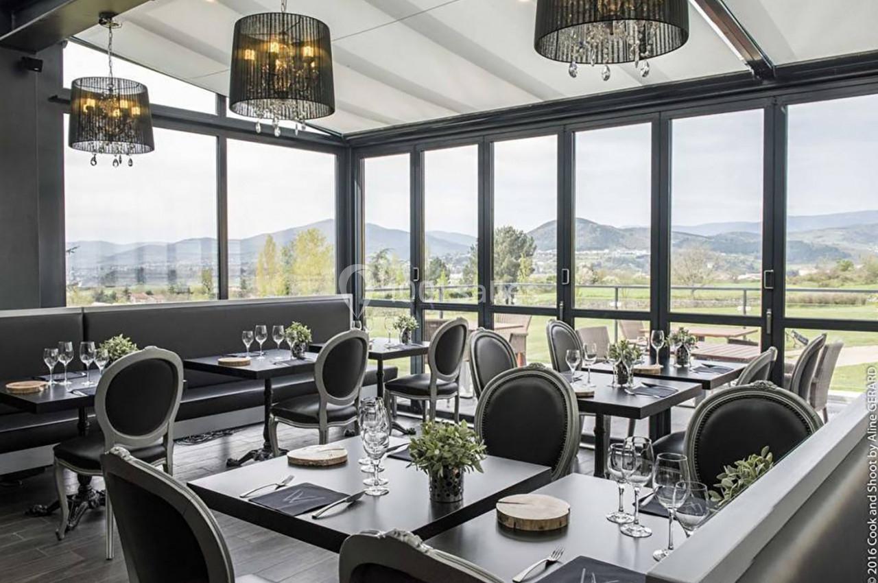 Salle de restaurant lumineuse avec baies vitrées donnant sur un paysage de collines, tables dressées avec élégance.