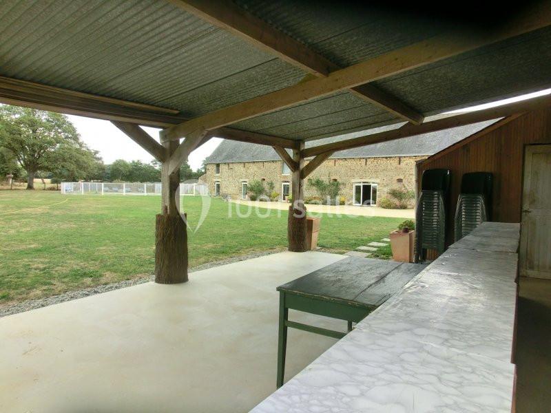 Vue d'une terrasse couverte avec un plan de travail en marbre, donnant sur un jardin et une maison en pierre.