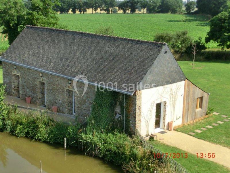 Bâtiment en pierre avec toit en ardoise, entouré de verdure, près d'un plan d'eau et d'un champ.