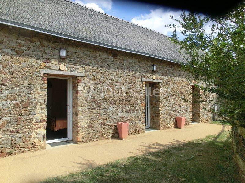 Façade en pierre d'une maison avec trois portes, pots de fleurs et allée en gravier, entourée de verdure.