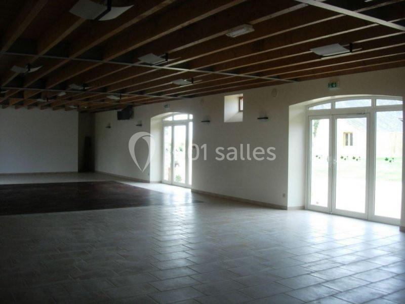 Salle vide avec sol carrelé, plafond en poutres apparentes, grandes fenêtres et portes vitrées laissant entrer la lumière…