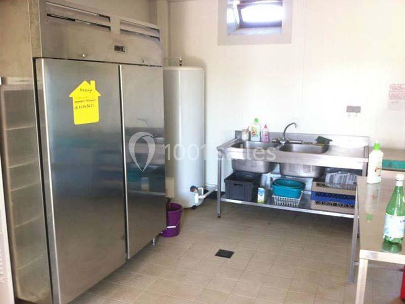 Cuisine professionnelle avec réfrigérateur en inox, évier double, produits d'entretien et sol carrelé beige.