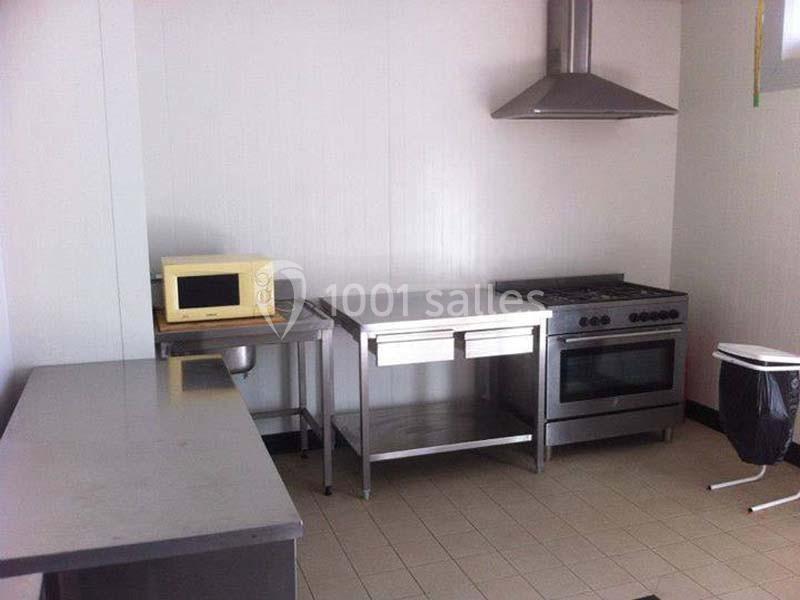 Cuisine équipée avec un four, une hotte, un micro-ondes, des plans de travail en inox et un sol carrelé beige.