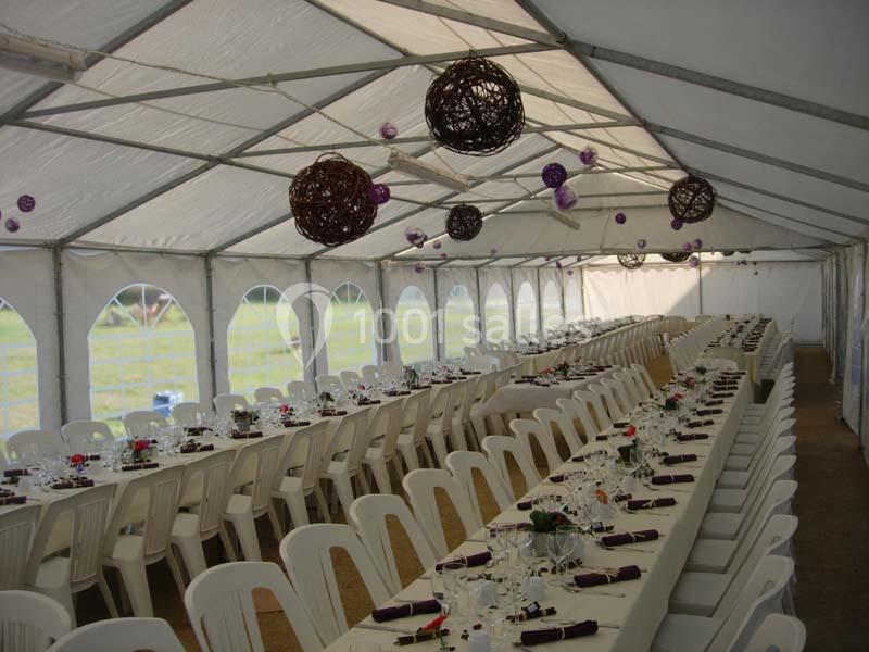 Intérieur d'une grande tente avec des tables alignées, décorées de nappes blanches, serviettes violettes et suspensions en…