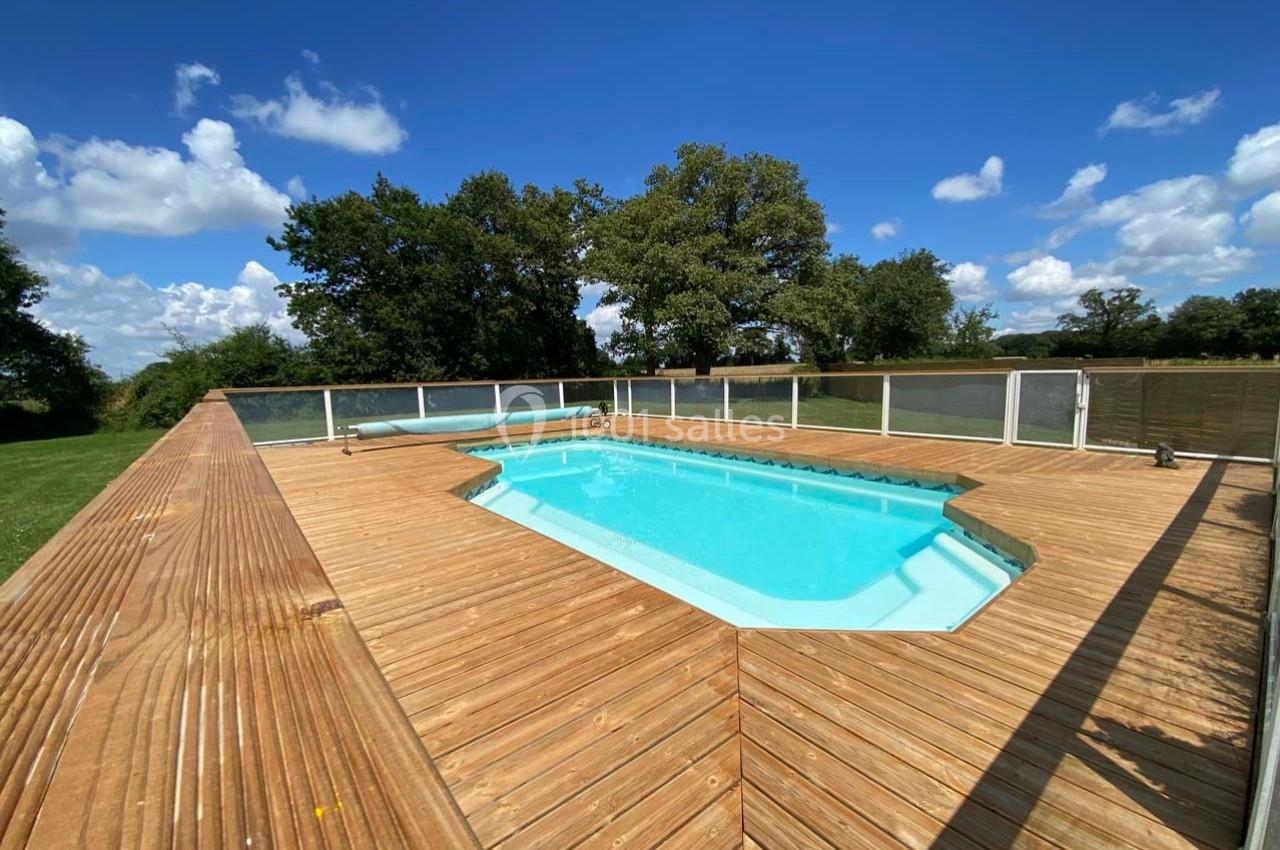 Piscine extérieure rectangulaire entourée d'une terrasse en bois, située dans un espace verdoyant sous un ciel bleu.